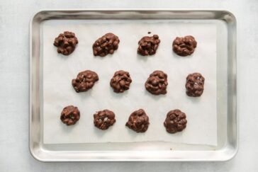 A baking tray lined with parchment paper holds twelve irregularly shaped chocolate clusters, each studded with what appear to be nuts or similar ingredients, against a light background.