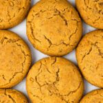 Close-up of round, golden-brown pumpkin cookies with cracked surfaces, arranged in a grid pattern on a light-colored background.