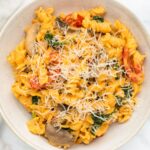 A bowl of creamy pasta topped with grated cheese. The pasta includes ingredients like sliced mushrooms, spinach, and cherry tomatoes, all coated in a rich sauce. The dish is placed in a neutral-colored bowl on a light surface.
