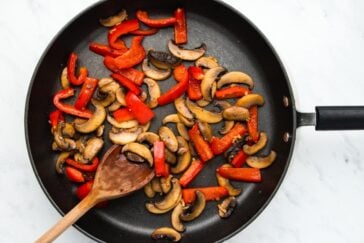 A frying pan filled with sautéed sliced mushrooms and red bell pepper strips. A wooden spoon rests on the edge of the pan, which sits on a light gray marble surface.