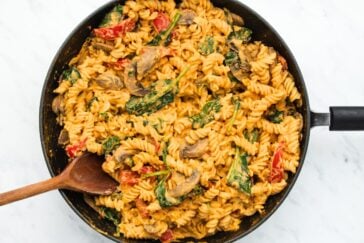 A skillet filled with creamy pasta, featuring fusilli noodles, mushrooms, spinach, and sliced red bell peppers. A wooden spoon rests inside, ready to serve the colorful and hearty dish.