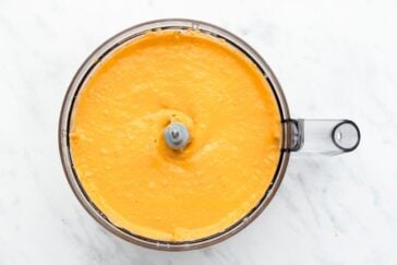 A food processor filled with smooth, creamy orange puree sits on a white marble surface.