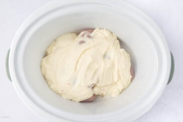 A white slow cooker contains raw chicken breasts covered in a thick, creamy mixture, possibly a sauce or marinade, featuring a pale yellow color. The cooker is on a white surface.