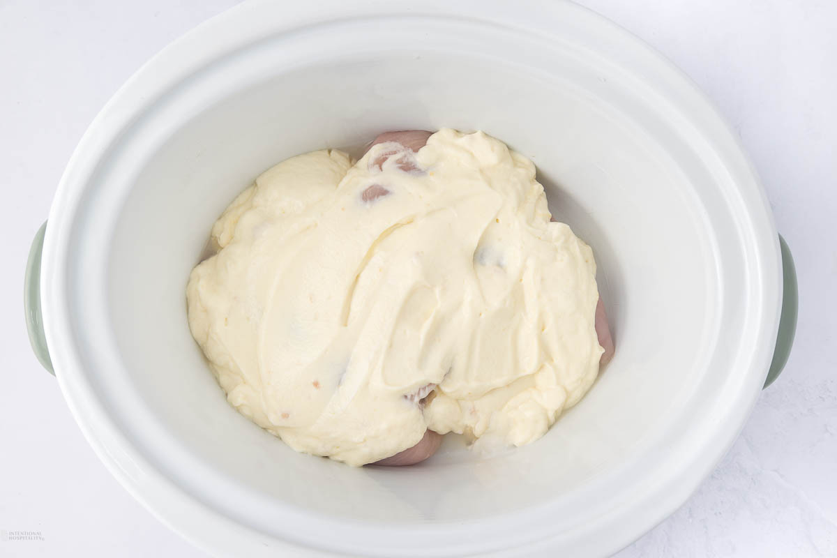 A white slow cooker contains raw chicken breasts covered in a thick, creamy mixture, possibly a sauce or marinade, featuring a pale yellow color. The cooker is on a white surface.