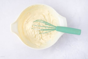 A white mixing bowl containing creamy batter with a green-handled whisk resting on the side. The batter appears smooth and well-mixed, set against a light gray background.