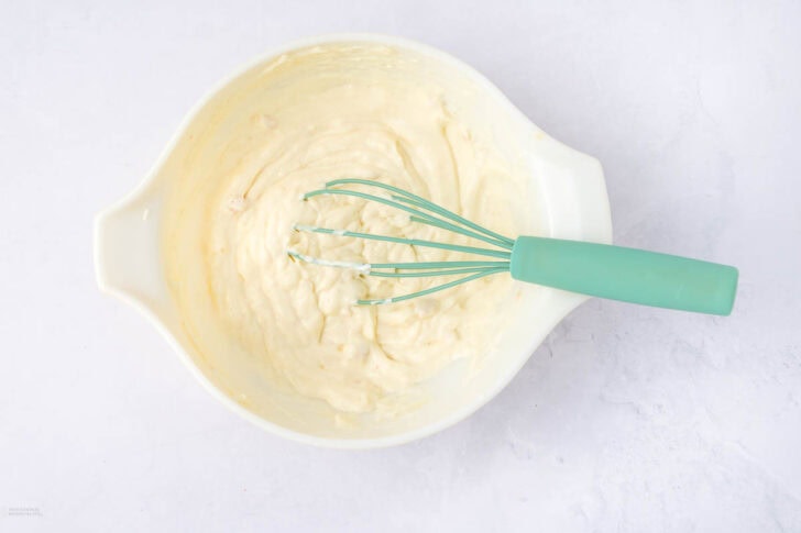 A white mixing bowl containing creamy batter with a green-handled whisk resting on the side. The batter appears smooth and well-mixed, set against a light gray background.