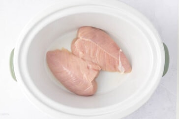 Two raw chicken breasts placed side by side in a white oval slow cooker. The cooker is empty, showing a smooth interior and green handles on the sides.