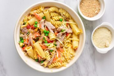 A bowl filled with pasta, mixed with pieces of tuna, peas, diced tomatoes, and shredded cheese. Next to it are two bowls: one with breadcrumbs and the other with grated cheese, against a marble countertop background.