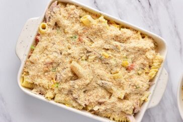 A casserole dish filled with a pasta bake topped with breadcrumbs. The baked pasta includes visible pieces of vegetables and shredded meat. The white dish rests on a marble countertop.