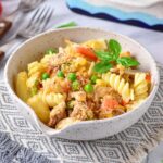 A ceramic bowl filled with fusilli pasta, peas, chunks of chicken, tomato pieces, and topped with breadcrumbs. Fresh basil leaves garnish the dish. The bowl is set on a patterned cloth with cutlery nearby.