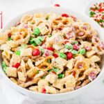 A bowl filled with a festive snack mix containing cereal squares, pretzels, red and green candies, and sprinkles, all coated in a white glaze.