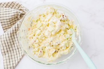 A bowl of creamy potato salad with chopped potatoes and vegetables, sitting on a white marble surface. A light blue spoon is in the bowl, and a checkered cloth is placed nearby.