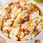A bowl of creamy salad topped with chopped chocolate bars and a drizzle of caramel sauce.