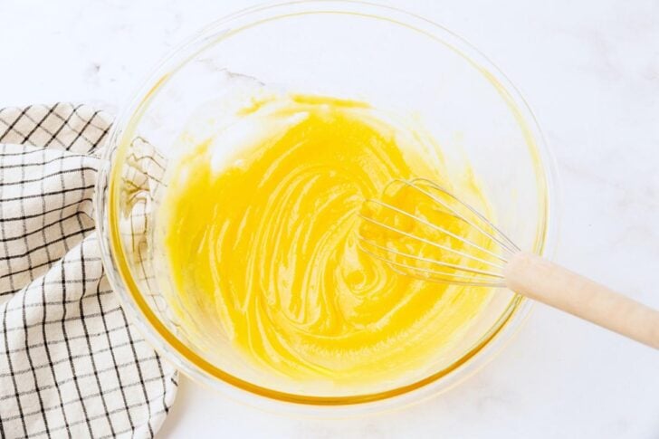 A glass bowl containing a smooth, yellow mixture, likely a batter, is being stirred with a whisk. A white and black checkered cloth is placed beside the bowl on a light-colored surface.