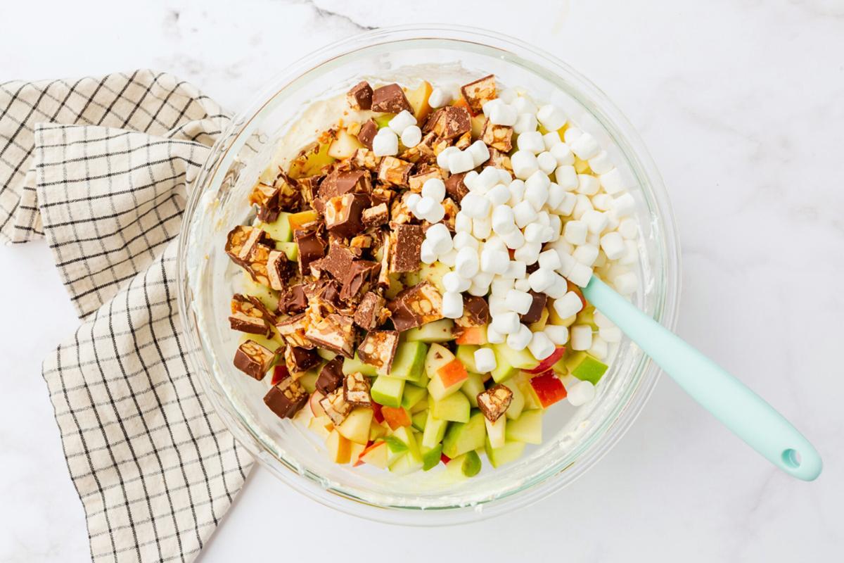 A glass bowl filled with chopped apples, snickers pieces, and mini marshmallows, with a creamy base. A light blue spatula is in the bowl. A black and white checkered cloth is beside the bowl on a marble surface.
