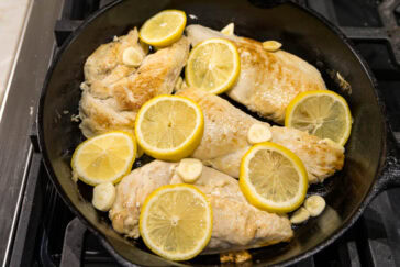 Sizzling chicken breasts in a cast iron skillet, topped with lemon slices and garlic cloves. The dish is cooking on a stovetop, with the lemons arranged over the lightly browned chicken.
