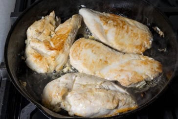 Four pieces of chicken breast being cooked in a sizzling black skillet, showing a golden-brown sear. Steam rises from the hot pan.