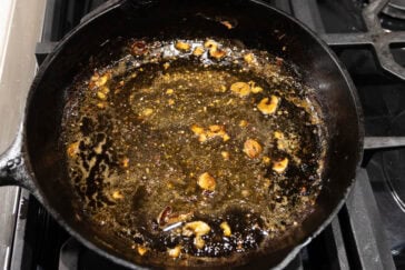 A black cast iron skillet on a stove top, containing remnants of browned, crispy garlic and leftover oil. The surface has a textured, seasoned appearance.