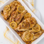 A white rectangular dish filled with baked chicken breasts, garnished with lemon slices and seasoned with spices. The dish is placed on a yellow and white checkered towel.