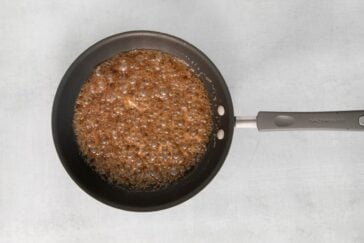 A non-stick frying pan on a gray surface, containing bubbling brown caramel sauce. The sauce covers the bottom of the pan, appearing glossy with small bubbles throughout.