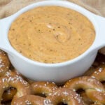 A white bowl brimming with creamy pretzel cheese dip is surrounded by soft, salted pretzels. These golden treats are artfully arranged on a wooden board, set against a backdrop of a beige cloth.