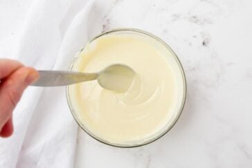 A hand holds a metal spoon over a glass bowl filled with creamy, white yogurt, set on a marble countertop. A folded white cloth is partially visible to the side.