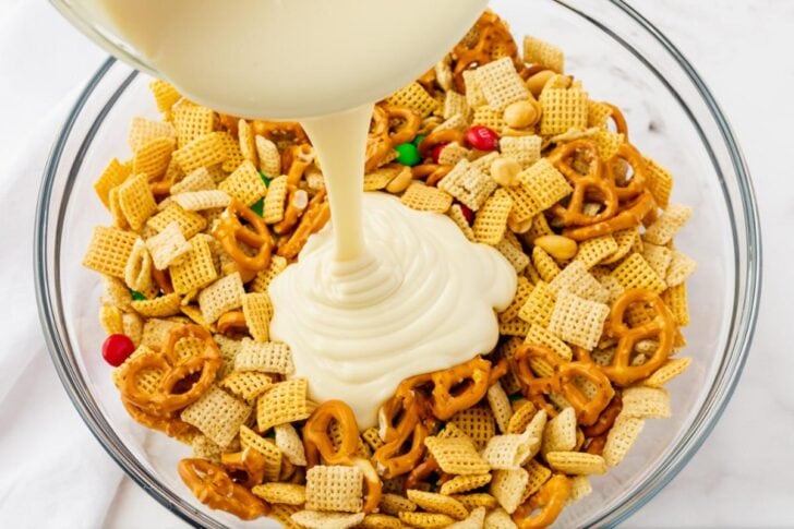 A bowl filled with Chex cereal, pretzels, peanuts, and colorful chocolate candies is being covered with a stream of white melted candy or chocolate coating. The ingredients are displayed on a marble surface.