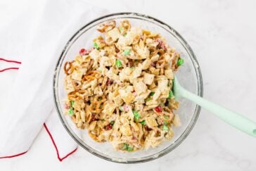 A glass bowl filled with a snack mix, including pretzels, cereal squares, colorful candies, and coated in a white mixture. A light green spatula is resting inside. The background is a white surface with a red-trimmed cloth nearby.