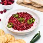 A bowl of cranberry salsa dip topped with sliced jalapeños, surrounded by sliced bread, a small bowl of cranberries, and crackers. A fresh jalapeño is also placed nearby on a white surface.