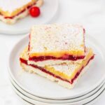 Two slices of lemon raspberry bars dusted with powdered sugar are stacked on a white plate. Another plate with a single bar and a raspberry is visible in the background.