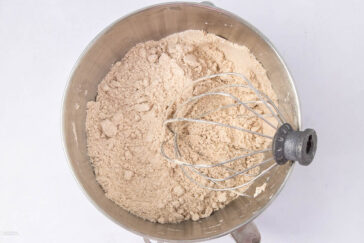 A metallic mixing bowl filled with dry ingredients and a whisk attachment placed inside. The light brown mixture appears to be flour-based, possibly for baking. The surface around the bowl is plain and clean.