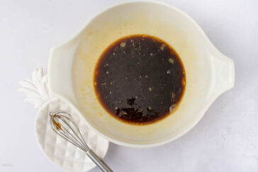A white mixing bowl containing a dark liquid marinade. Next to it is a small white dish with a metal whisk. The background is a light-colored countertop.