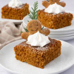 A square slice of gingerbread cake topped with whipped cream, a small gingerbread man cookie, and a sprig of greenery. The cake is dusted with powdered sugar and set on a white plate with additional slices in the background.