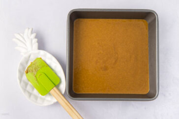 A square baking pan filled with brown batter is placed on a white surface. Beside it, a green spatula with batter rests on a white, pineapple-shaped dish.