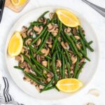 A plate of cooked green beans and sliced mushrooms garnished with lemon wedges. The dish is served on a white plate on a marble surface, with a striped napkin partially visible at the edge.