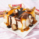 A slice of cake with caramel and chocolate drizzles, topped with pieces of chocolate bar, on a white plate. Wrapped mini chocolate bars are in the background on a pink and white striped surface.