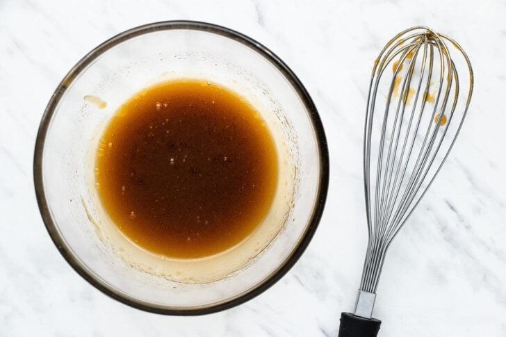 A glass bowl filled with a brown liquid mixture sits on a marble surface, next to a metal whisk with some mixture droplets on it.