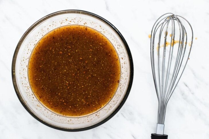 A clear glass bowl filled with brown liquid and spices on a marble surface. A metal whisk with a black handle lies beside the bowl, with traces of the mixture on the whisk.