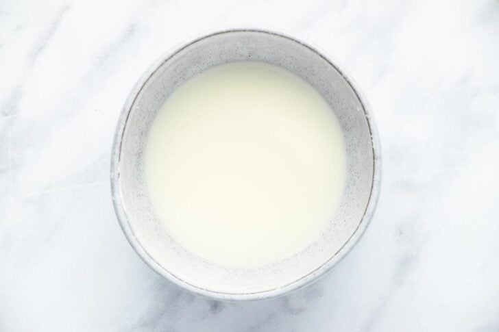 A bowl of milk is placed on a white marble surface. The bowl is simple and round with a smooth texture, filled to the brim with creamy milk. The background is light, enhancing the clarity and freshness of the scene.