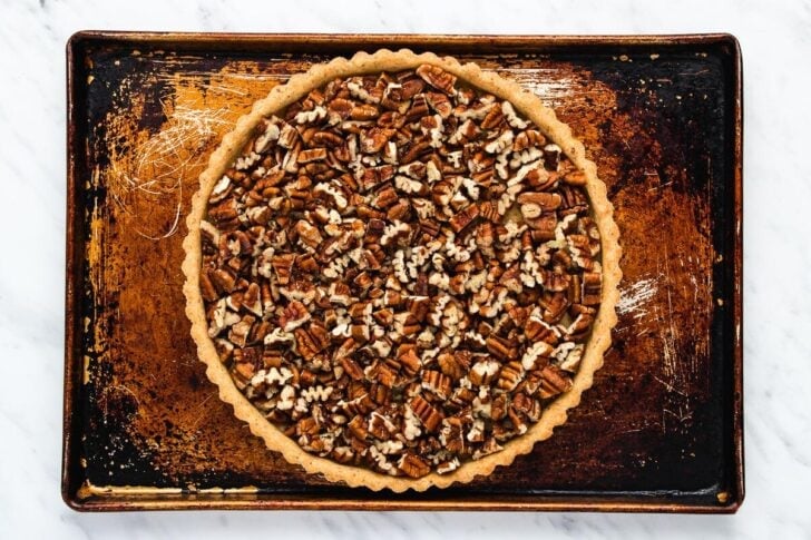 A pecan pie on a baking sheet, viewed from above. The pie crust is filled with a generous layer of whole and chopped pecans, contrasting with the dark, worn surface of the baking sheet beneath it.
