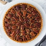 A freshly baked pecan pie with a golden crust, filled with whole pecans arranged neatly in a circular pattern, sits on a white plate. The pie is placed on a marble countertop.