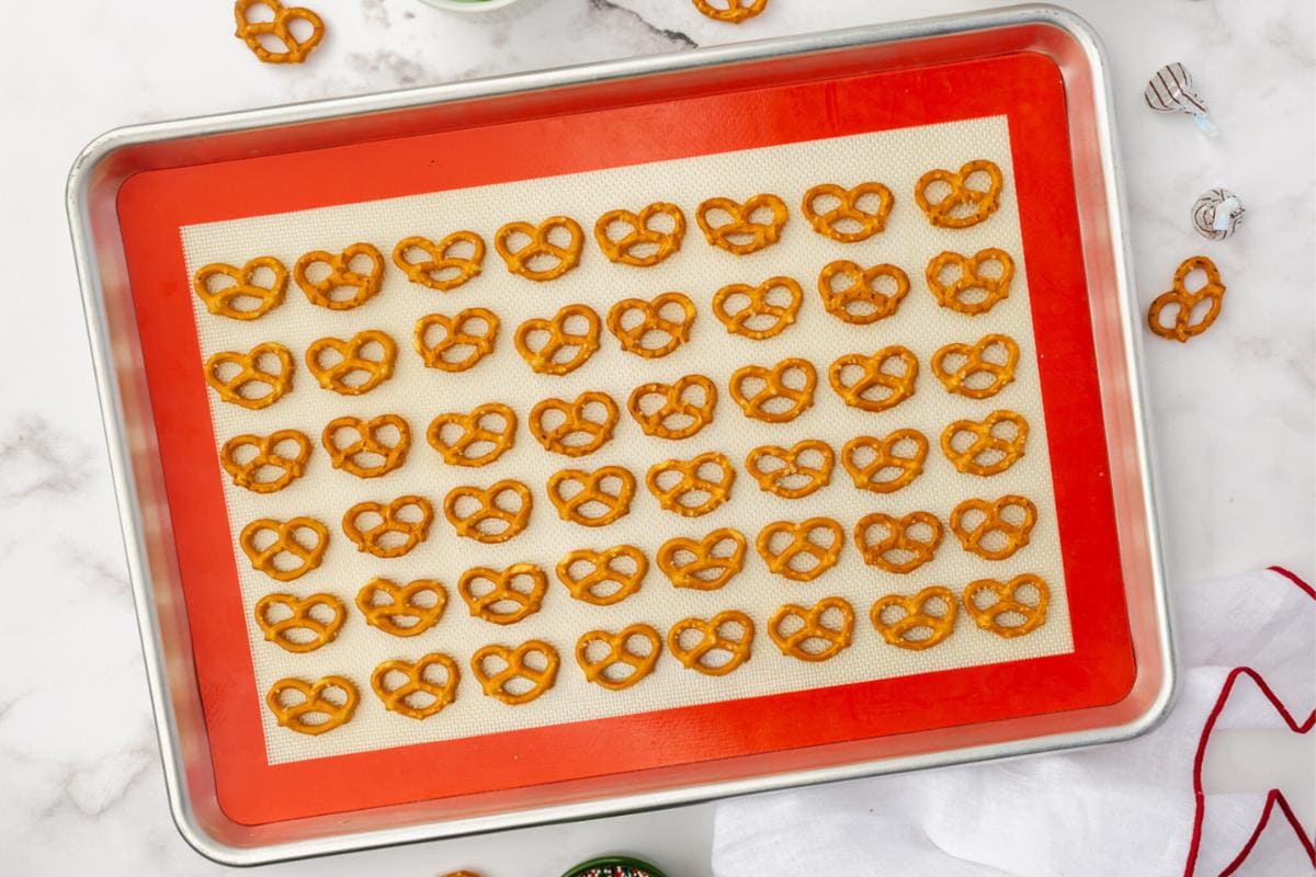 A baking tray lined with a silicone mat is filled with rows of small pretzels, arranged neatly. A red kitchen towel and a few wrapped chocolate candies are nearby on a marble countertop.