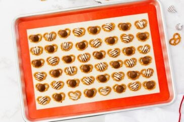 A baking tray lined with parchment paper holds rows of pretzels topped with melted chocolate and white chocolate drizzle. The pretzels are evenly spaced and neatly organized.
