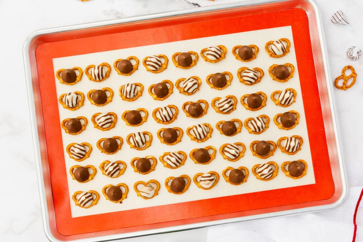 A baking tray lined with parchment paper holds rows of pretzels topped with melted chocolate and white chocolate drizzle. The pretzels are evenly spaced and neatly organized.