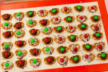 A tray of festive pretzel treats topped with melted chocolate, red and green candies, and sprinkles. Arranged in neat rows on a red background, the colorful snacks evoke a holiday theme.