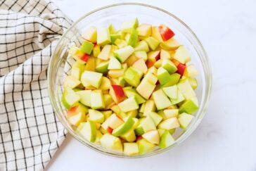 A glass bowl filled with diced red and green apples on a marble surface. A white cloth with black checkered pattern rests beside the bowl.