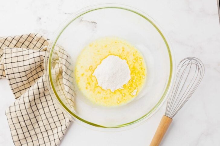 A glass bowl with butter and a mound of flour sits on a marble countertop next to a checkered cloth. A whisk is placed beside the bowl.
