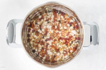 A stainless steel pot filled with a variety of dried beans and lentils soaking in water. The colorful mixture includes beans of different shapes and sizes, floating in the clear liquid. The pot is placed on a light surface.
