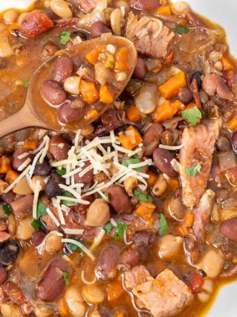 A bowl of hearty bean stew with chunks of meat, diced carrots, and a wooden spoon. The dish is topped with shredded cheese and garnished with herbs. Crackers are placed beside the bowl on a wooden surface.