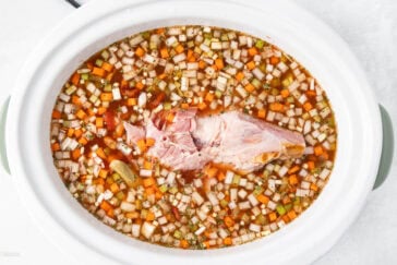 A white slow cooker filled with a broth containing diced carrots, onions, celery, and seasonings, with a large piece of ham in the center.